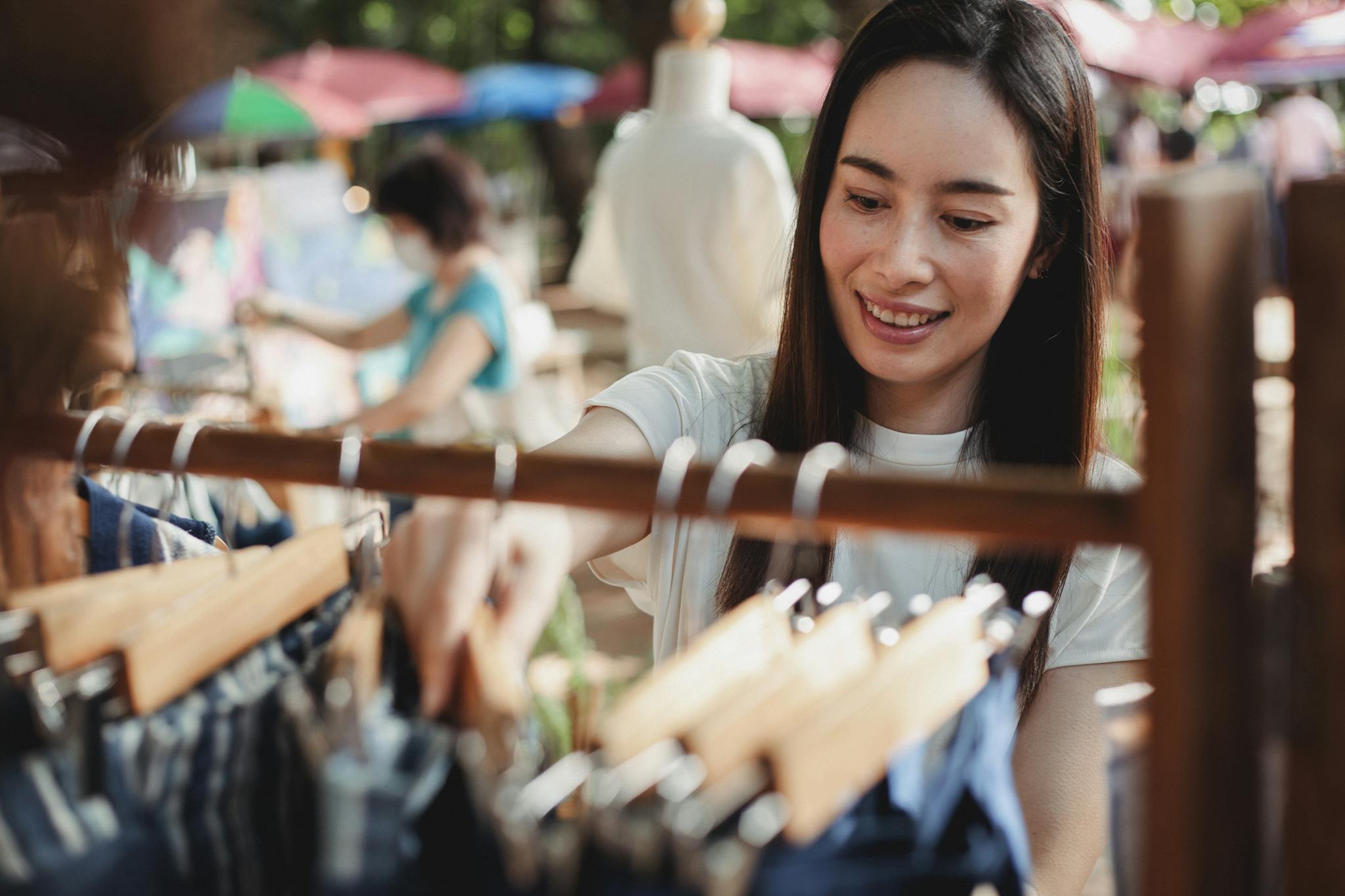 Asian woman enjoying shopping at an outdoor market, browsing through clothes hangers with a smile.