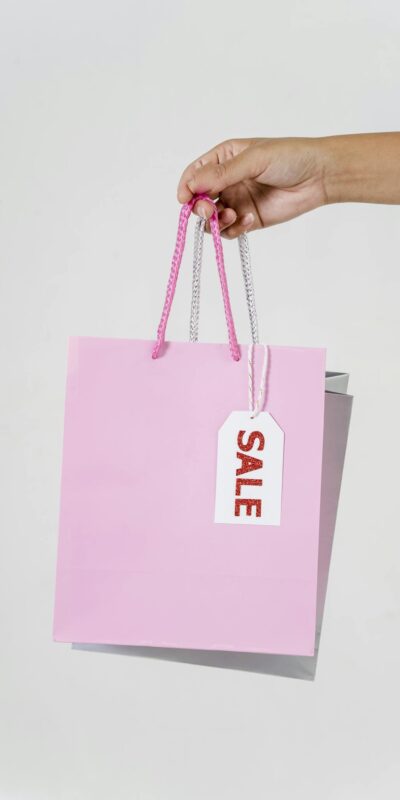 Close-up of a hand holding a pink shopping bag with a sale tag against a white background.