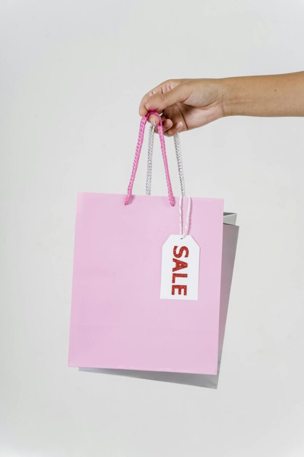 Close-up of a hand holding a pink shopping bag with a sale tag against a white background.